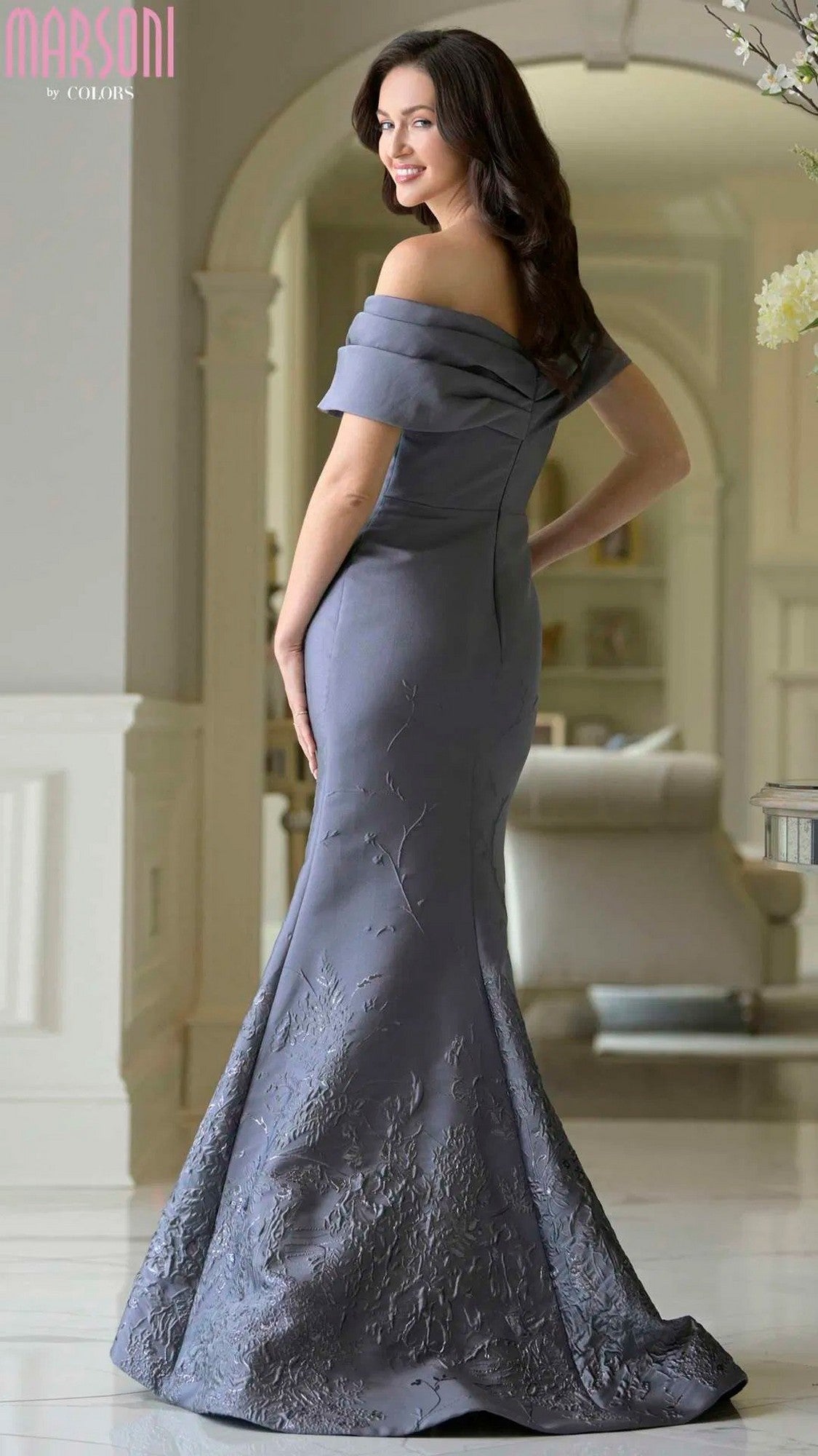 Woman stands indoors in a Marsoni MV1378 long formal dress—an asymmetric off-shoulder, floor-length grey mermaid gown with floral embroidery—smiling over her shoulder.